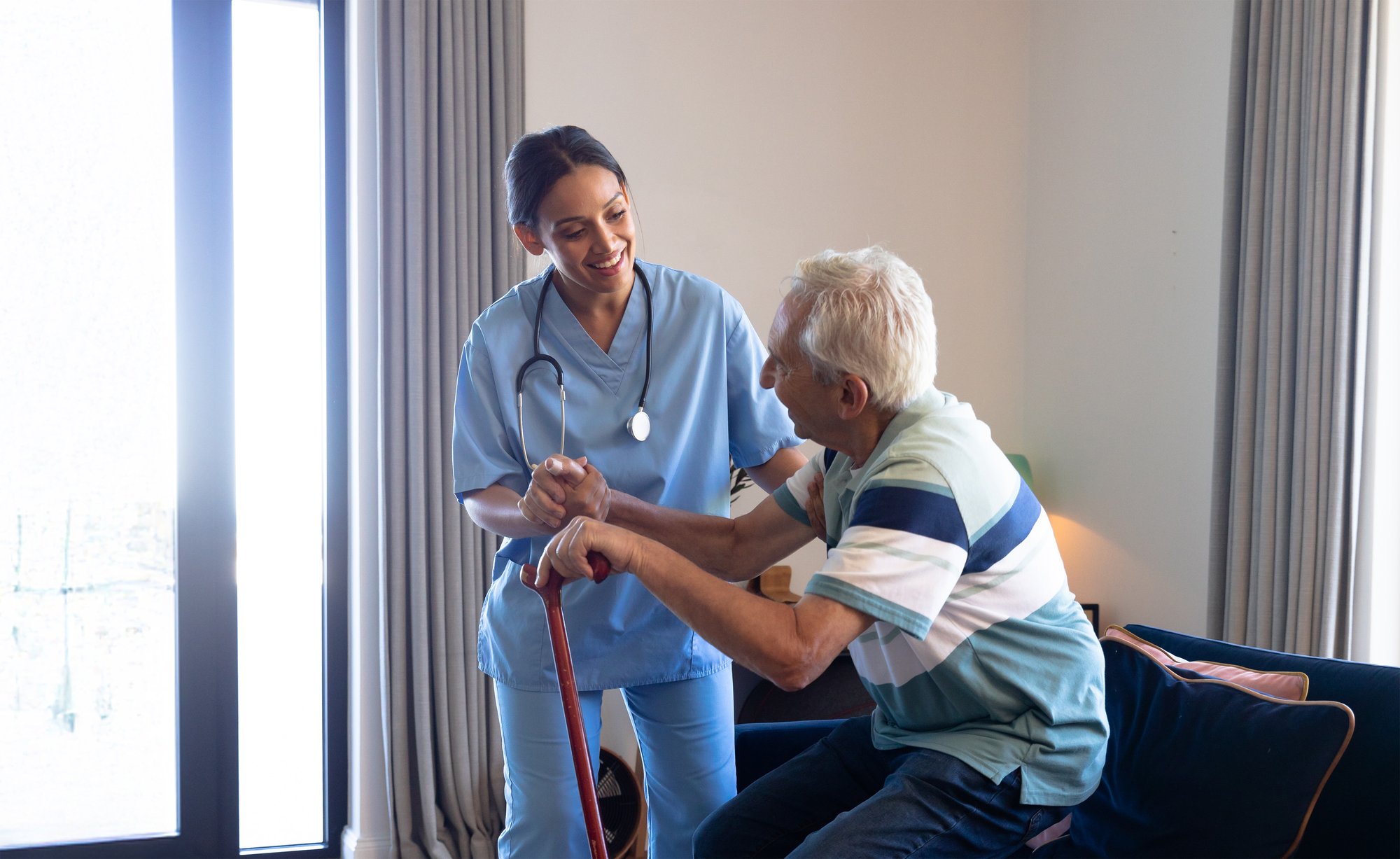 biracial-female-physiotherapist-assisting-caucasian-senior-man-getting-up-from-sofa-home-bearb